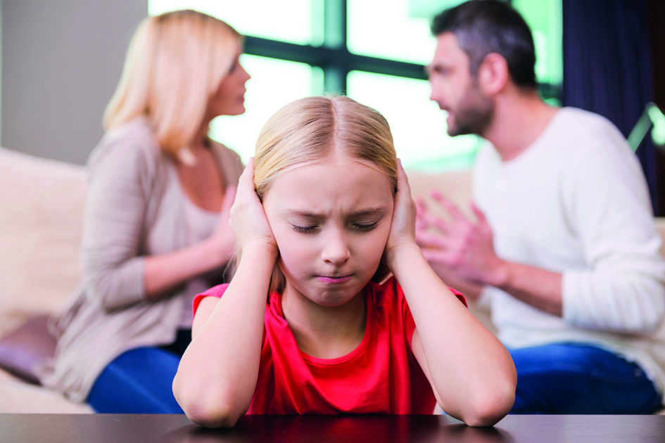 Please stop fighting! Depressed little girl leaning at the table and covering ear with hands while her parents shouting at each other in the background
